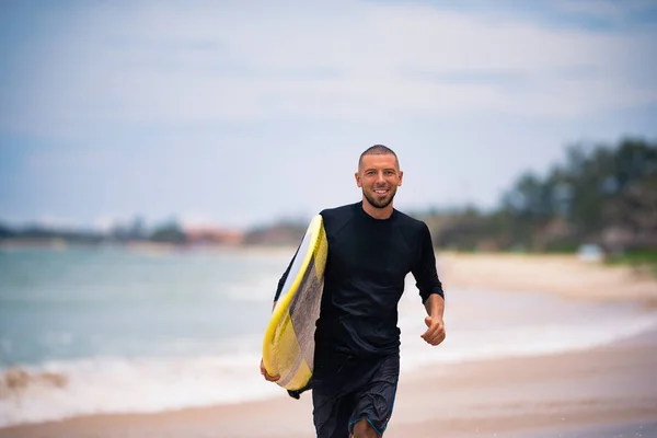 Sorridente attraente giovane surfista in esecuzione con tavola da surf gialla sulla spiaggia soleggiata. Vacanze estive outdoor stile di vita attivo. — Foto Stock