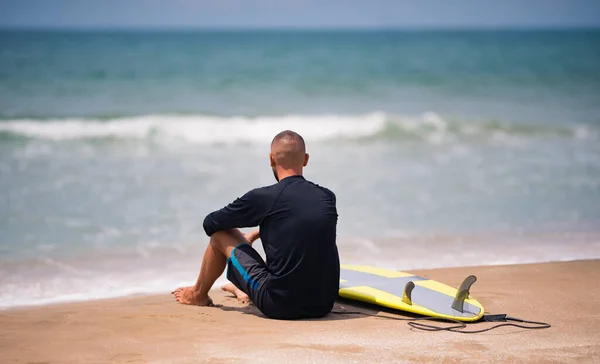 Junger Surfer mit Surfbrett sitzt allein am Strand und blickt aufs Meer und wartet auf Wellen. — Stockfoto