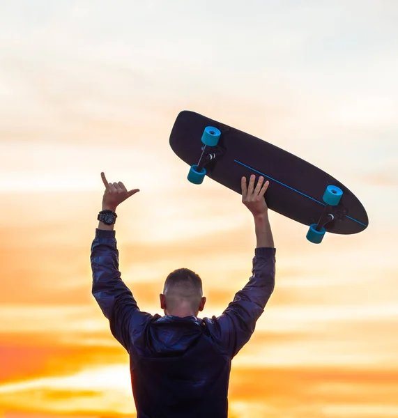 Мужчина наслаждается беззаботной жизнью на закате. Держит скейтборд и показывает знак шака. — стоковое фото