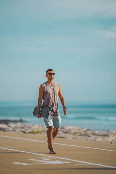 Uomo spensierato e sportivo che si gode la vita e cammina vicino all'oceano con il longboard in mano al tramonto. Concetto attività estive. — Foto Stock