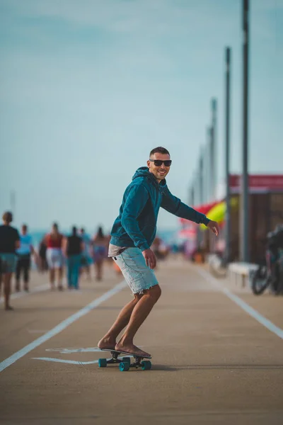 Gelukkig zorgeloze en sportieve jongeman in een capuchon en zonnebril rijdt op een surfplank. Zomer leuke activiteiten concept. — Stockfoto