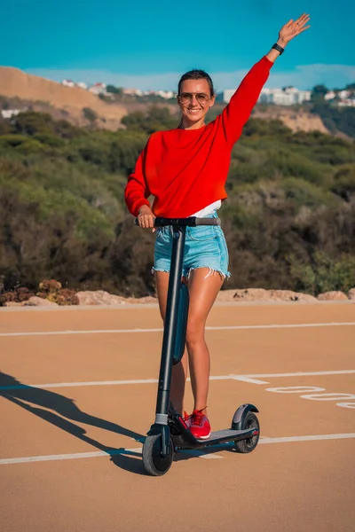 빨간 후드를 입은 행복 한 젊고 스포츠적 인 여자와 전기 스쿠터를 들고 서 있는 파란 짧은 청바지를 입은 사람. 여름 야외 활동. — 스톡 사진