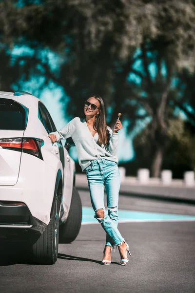Schattige pasvorm brunette vrouw in stijlvolle kleding met lolly staat in de buurt van haar nieuwe witte electro auto in zonnige zomerdag. Succesvolle jonge zakenvrouw. — Stockfoto