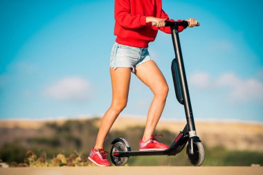 Kırmızı kapüşonlu ve mavi kot şortlu genç kız yolda elektrikli scooter gidonu tutuyor. Yazın açık hava aktiviteleri.