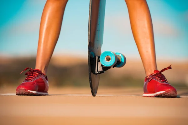 Mujer deportiva con hermosas piernas bronceadas en elegantes zapatillas rojas sostiene el patín de surf entre sus piernas en el día de verano brillante. Actividades al aire libre de verano. Cultura callejera. — Foto de Stock