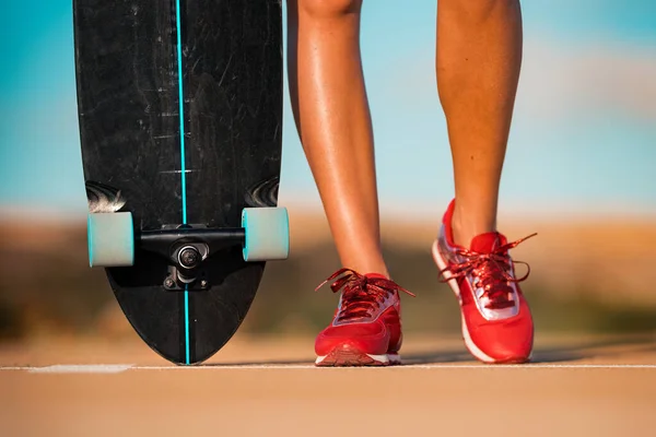 Fit vrouw met mooie gebruinde benen in stijlvolle rode sneakers houdt zwart longboard op glanzende zomerdag. Buitenactiviteiten in de zomer. Straatcultuur. — Stockfoto