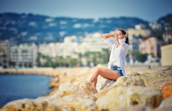 Carino sorridente donna spensierata in camicia bianca e pantaloncini blu è seduto su pietre vicino all'oceano. — Foto Stock