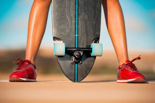 Foto de primer plano del patinador de mujer en forma y deportivo con hermosas piernas sexy en elegantes zapatillas rojas sostiene longboard entre sus piernas en el día de verano brillante. Actividades al aire libre. Cultura callejera. — Foto de Stock