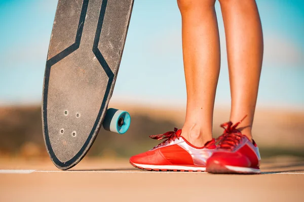 Güzel bronzlaşmış bacakları ve şık kırmızı spor ayakkabıları olan kadın kaykaycı parlak yaz gününde sörf tahtası tutuyor. Yaz zamanı açık hava aktiviteleri. Sokak kültürü. — Stok fotoğraf