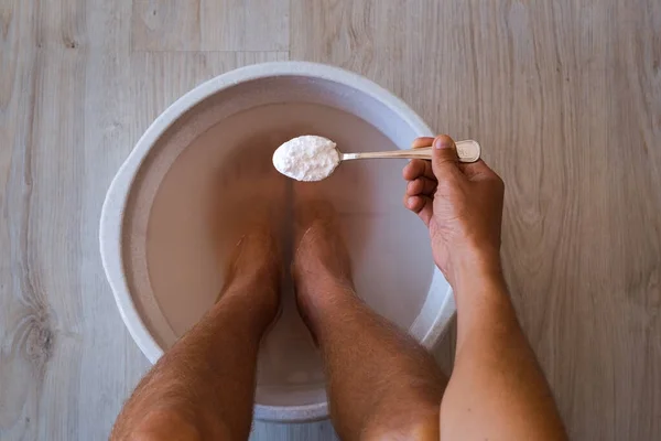 Seorang pria menaruh sendok baking soda di kamar mandi dengan air panas untuk kakinya. Mandi buatan sendiri rendam untuk kulit kaki kering — Stok Foto