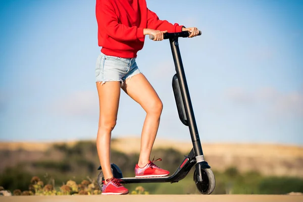 Happy woman in red hoodie and blue short jeans standing with rented electric scooter. New generation, electric transport, ecology, ecological transport. The concept of a healthy and green lifestyle — Stock Photo, Image