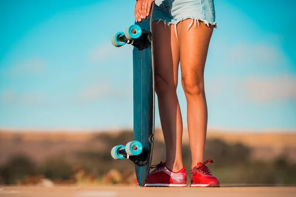 Подходящая женщина с тощими загорелыми ногами в стильных джинсовых шортах держит скейтборд. Здоровое увлечение на открытом воздухе — стоковое фото