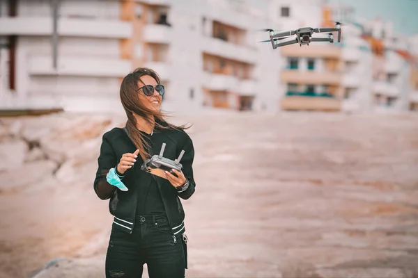 Ziemlich moderne lächelnde Pilotin in dunkel-stylischer Kleidung fliegt Drohne bei starkem Wind in der Stadt. Drohne mit Genehmigung fliegen. Hobby im Freien. — Stockfoto