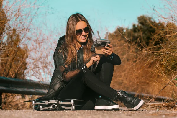 Giovane donna moderna in eleganti abiti scuri e occhiali da sole che si prepara a iniziare a volare drone al tramonto o all'alba. — Foto Stock