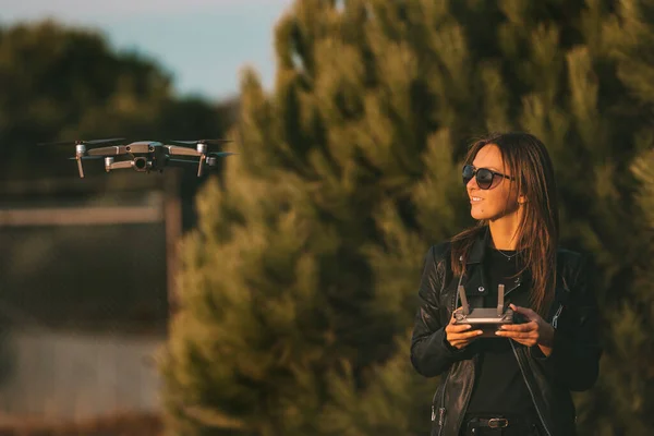 Young modern smiling woman pilot in dark stylish clothes operating the drone by remote control. Flying drone in forest at sunset. Female launches a drone for flight in the evening.