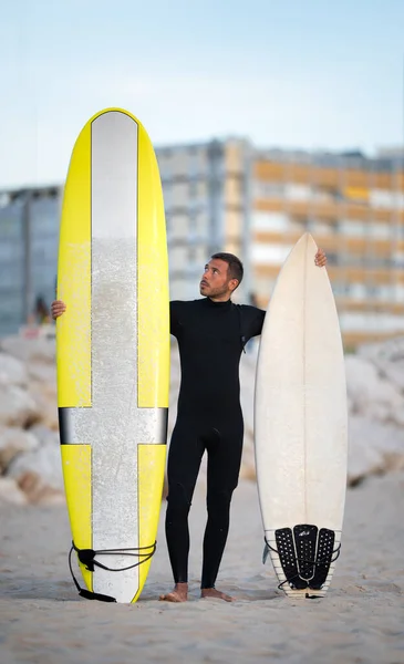 Πορτρέτο του νεαρού σέρφερ σε μαύρο ζίπλες surfing wetsuit προσπαθεί να επιλέξει longboard ή shortboard στην Costa Da Caparica, Πορτογαλία — Φωτογραφία Αρχείου