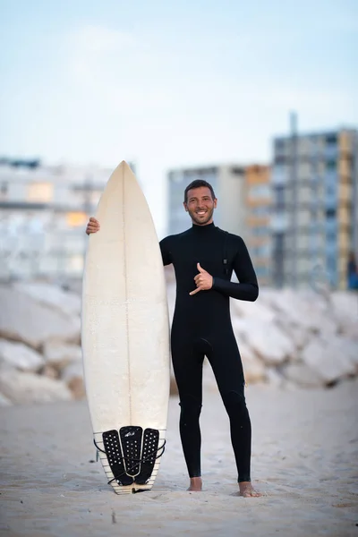 Junger lächelnder Surfer im schwarzen, reißverschlussfreien Surfanzug mit Shortboard mit Shaka-Zeichen in Costa Da Caparica, Portugal — Stockfoto