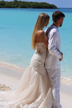 Gelinlik giymiş gelin ve beyaz takım elbiseli damat okyanus sahilinde sarılıyorlar.