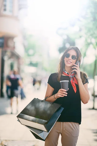 手持纸袋和咖啡的女性肖像在购物的同时使用智能手机 — 图库照片