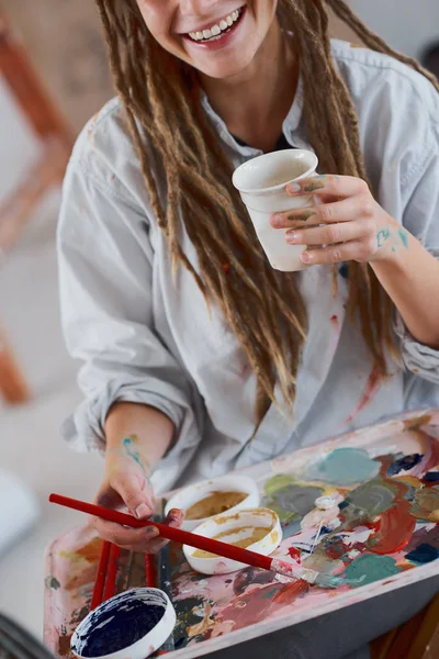 艺术女孩喝咖啡 而坐在演播室和绘画在画架上 — 图库照片