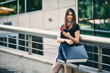 Alışveriş torbaları tutarak ve alışveriş merkezi önünde akıllı telefon kullanan kadın.