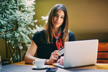 Kafede otururken alışveriş için kredi kartı ve dizüstü bilgisayar kullanan mutlu genç kadın.