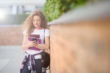 Akıllı telefon kullanarak ve kampüs içinde yürürken kitap tutan kız öğrenci.