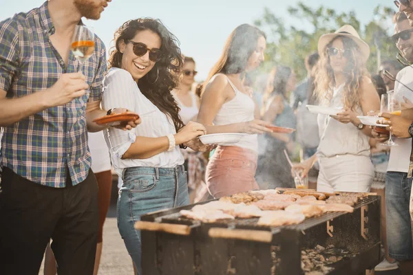 Group of people standing around grill, chatting, drinking and eating.