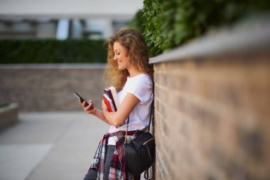 Akıllı telefon kullanarak ve kampüs içinde yürürken kitap tutan kız öğrenci.