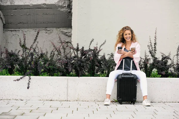 女旅行者使用智能手机 坐在墙上拿着咖啡 她旁边的行李 — 图库照片