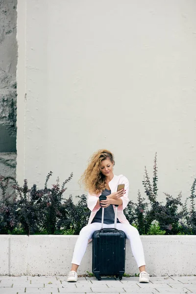 Kobiece Podróżnik Pomocą Smartfona Przytrzymując Kawy Siedząc Ścianie Bagażu Obok — Zdjęcie stockowe