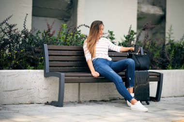 Otobüs istasyonunda bekleyen ve bankta oturan kadın. Bavulunu yanında. El kahve içinde.