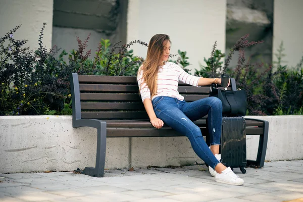 女人在车站等车 坐在长凳上 在她的行李旁边 在手咖啡去 — 图库照片