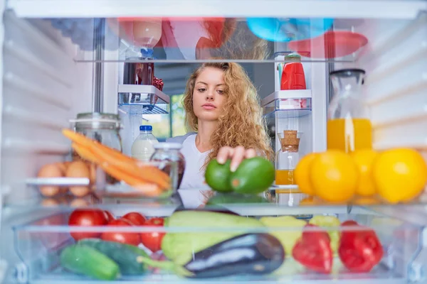 Woman standing in front of opened fridge and taking avocado. Fridge full of groceries.