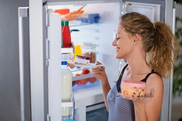 Woman taking cakes while standing in front of opened fridge.