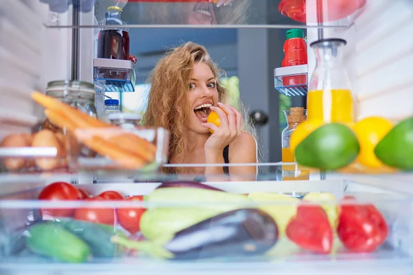 Woman biting fruit while standing in front of fridge full of groceries. Picture taken from the inside of fridge.