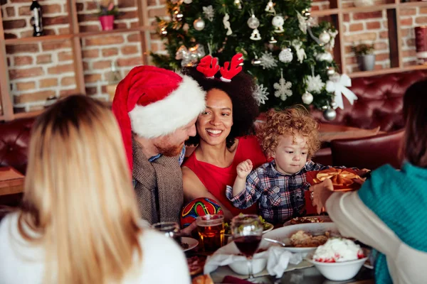 Friends giving gift to toddler while sitting at table. Christmas tree in background. Christmas holiday concept.