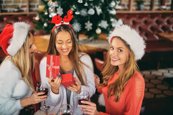 Friends giving gifts to each other and drinking wine while sitting at table. In background Christmas tree. Christmas holidays concept.
