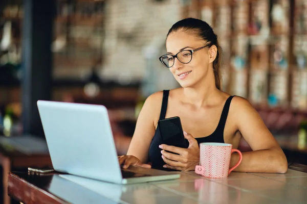 Woman using laptop and in other hand holding smart phone. On table mug with tea. Cafe interior.