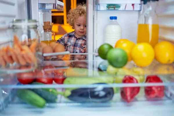 Toddler peeking in the fridge full of groceries and looking something to eat. Picture taken from the inside of fridge.
