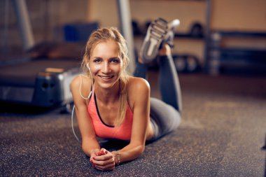 Spor salonunda yerde yatarken ve kameraya bakarak gülümseyen Genç beyaz bir kadın. Kulak kulaklık. Sağlıklı yaşam konsepti.