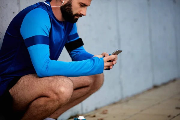 Young bearded man crouching and using smart phone. In background wall. Healthy lifestyle concept.