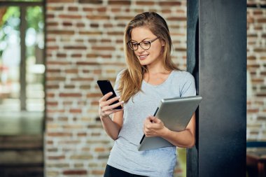 Duvara karşı dururken akıllı telefon kullanarak iş kadını. Dizüstü tutan diğer elinde.