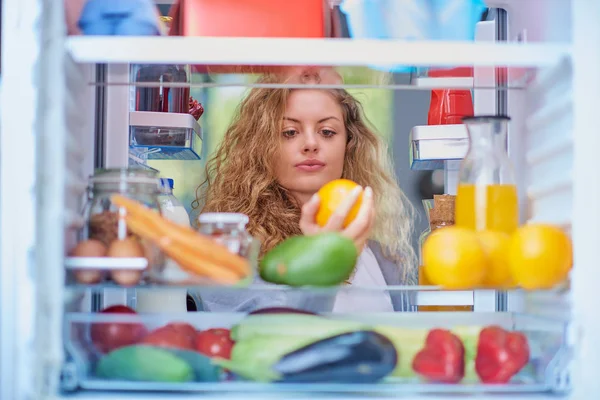 Woman standing in front of opened fridge and taking orange fruit. Fridge full of groceries. Picture taken from the iside of fridge.