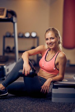 Spor salonu katta oturan ve şişe su ile tutan sportif kadın. Boyun kulaklık, spor giyim üzerine. Sağlıklı yaşam konsepti.