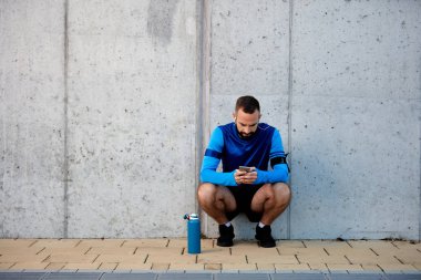 Genç adam çömelmiş ve akıllı telefon kullanarak sakallı. Onun yanında şişe su ile. Arka duvarda. Sağlıklı yaşam konsepti.
