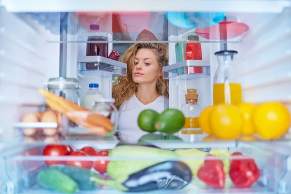 Woman standing in front of fridge full of groceries and looking something to eat. Picture taken from the inside of fridge.