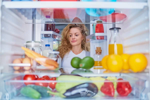Woman standing in front of fridge full of groceries and looking something to eat. Picture taken from the inside of fridge.
