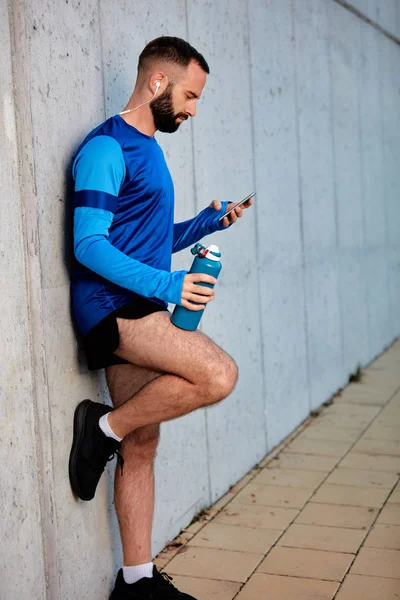 Corredor Masculino Apoyado Pared Utilizando Teléfono Inteligente Otra Botella Mano —  Fotos de Stock