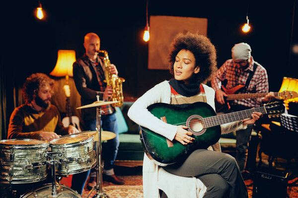 Смешанная расовая женщина играет на акустической гитаре и сидит со скрещенными ногами в домашней студии. На заднем плане остальные участники группы играют на барабанах, саксофоне и бас-гитаре
.